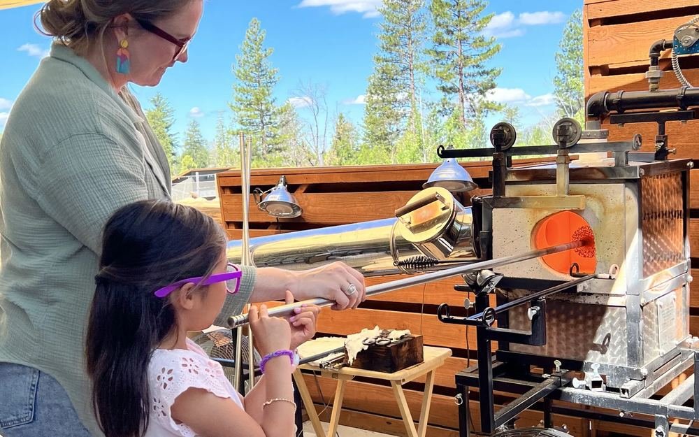 A young guest in safety glasses pulls her first gather of glowing glass from the furnace with an instructor beside her.