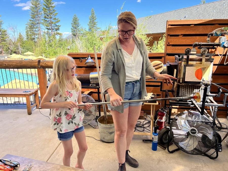 An instructor and a young guest hold a blowpipe together, a glowing gather at its end.