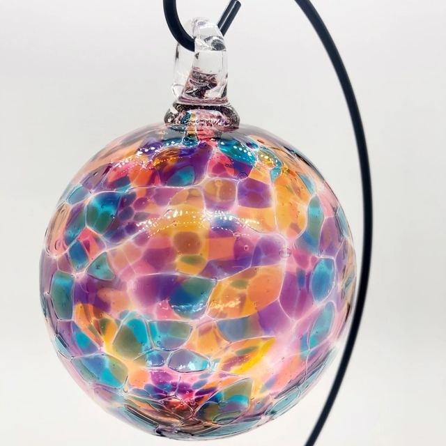 Round glass ornament in pink, teal, amber and violet hanging from a black stand.