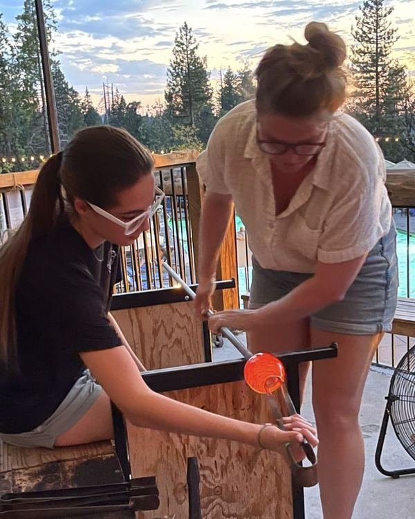 At dusk, an instructor guides a guest shaping a glowing orange bubble of glass at the bench, with pines behind.