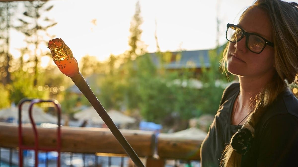 A glassblower at golden hour studies a glowing gather rolled in colored glass on the end of her pipe.